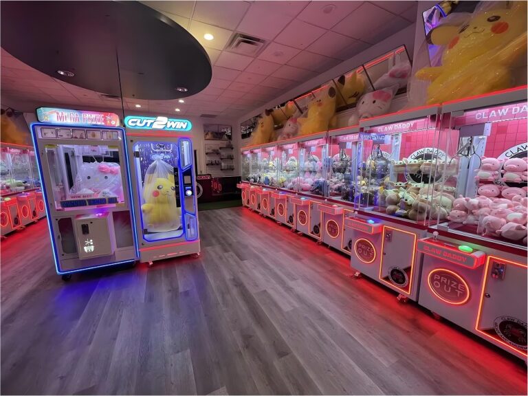 Interior de arcade con filas de máquinas de garra iluminadas por LED rojos y gabinetes con grandes premios, que demuestra una implementación en el mundo real y una configuración de alto tráfico.